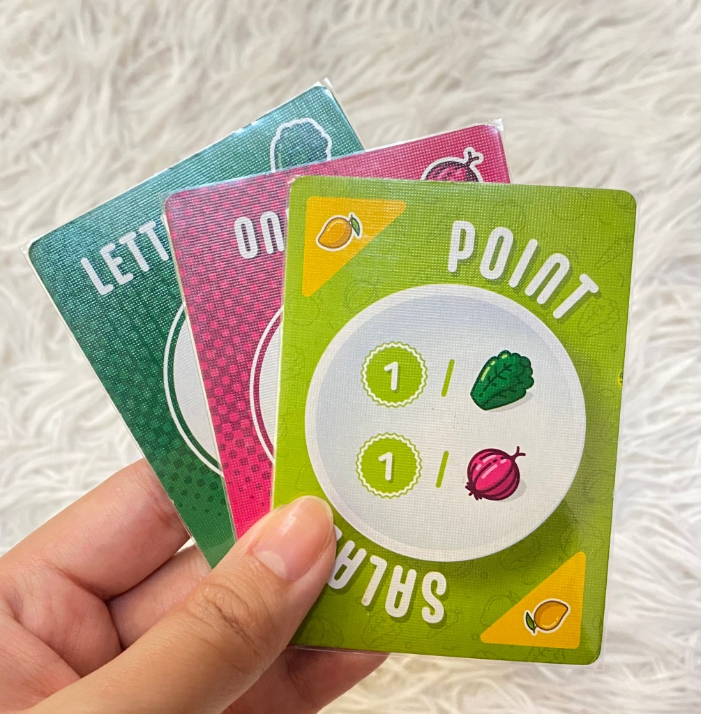 A hand holding Point Salad cards, featuring colorful vegetable illustrations.